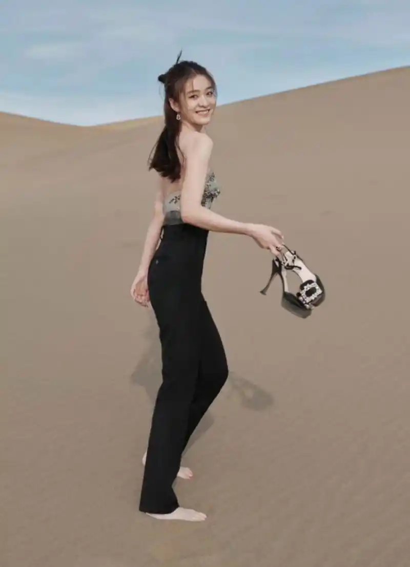 张雪迎在沙漠里参加活动,高跟鞋站不稳,用手提着拍照,回身看镜头甜笑